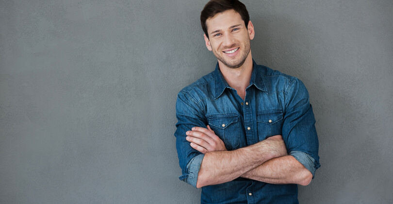 Picture of man standing with arms folded, representing the plastic surgery procedures available for males with Frontline Plastic Surgery, San Jose, Costa Rica.  The man is wearing a blue shirt and is smiling while facing the camera.