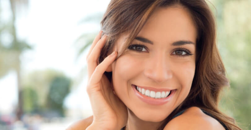 Close-up picture of a beautiful female patient, happy with the face lift and neck lift she had at Frontline Plastic Surgery in San Jose, Costa Rica.  The picture shows a woman with brown hair, holding her hand to the side of her face, and smiling while looking directly at the camera.